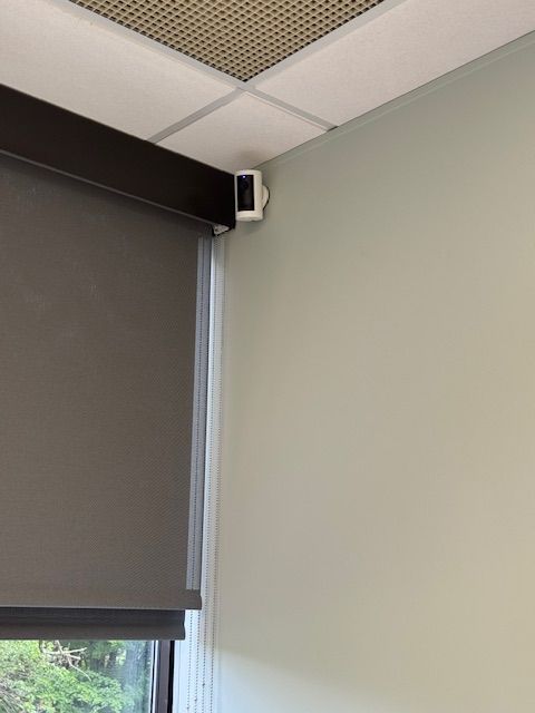 Security camera mounted in the corner of a room, next to a window with a grey blind.