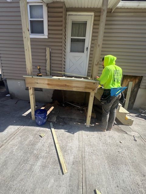 Person in neon green hoodie builds a small wooden porch outside a tan building with a white door.