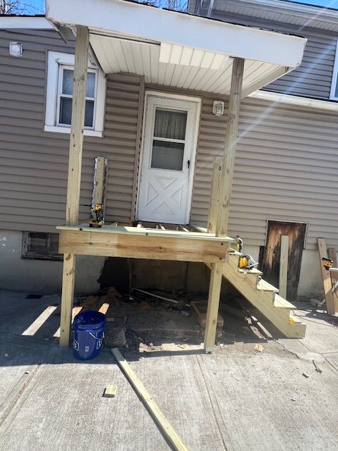 Wooden porch with steps leading to a back door of a building with light brown siding; blue bucket on the ground.
