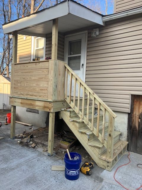 Wooden porch with stairs leading to a back door of a building. Blue bucket and tools at base.
