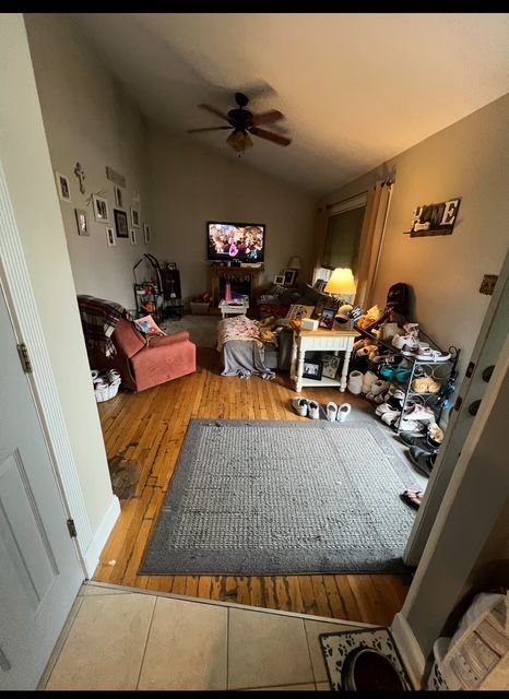Living room with cluttered furniture, rug on hardwood floor, TV on, and a ceiling fan.