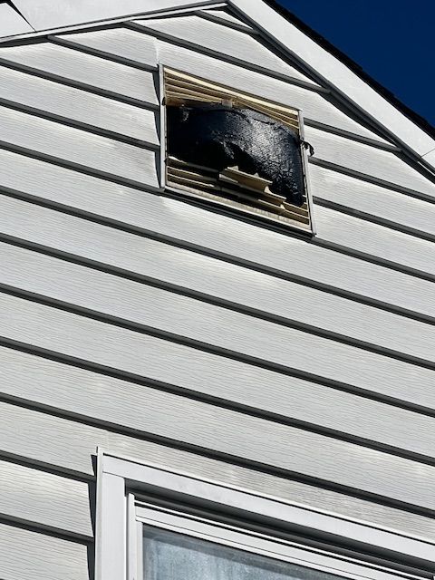 Damaged vent on a white vinyl-sided house; black, charred material visible.