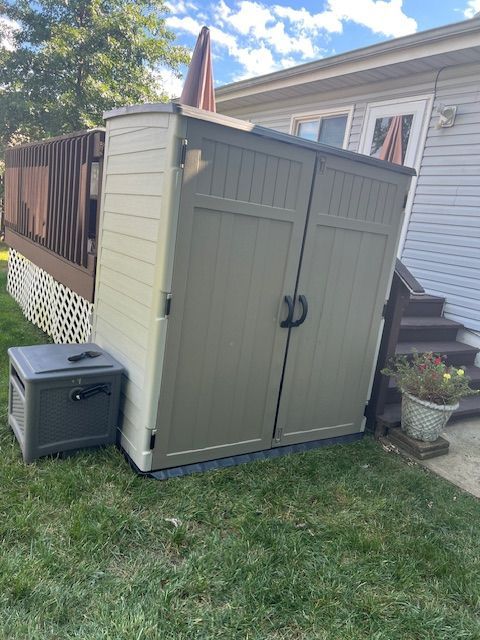 A tan storage shed next to a small gray box on a lawn. A deck and house are in the background.