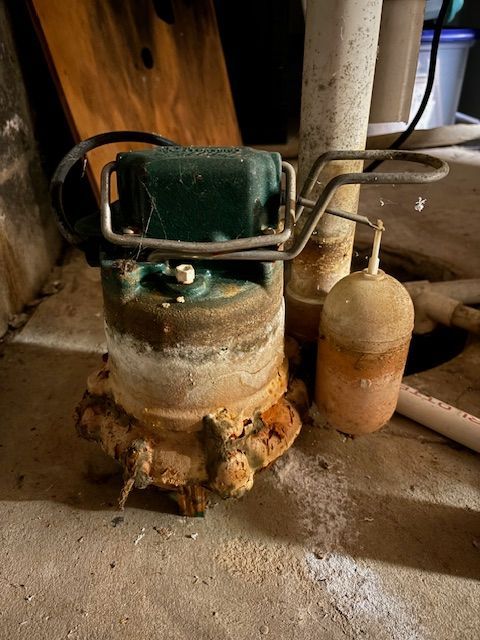 A rusty sump pump with a float switch, next to a white pipe, in a basement.