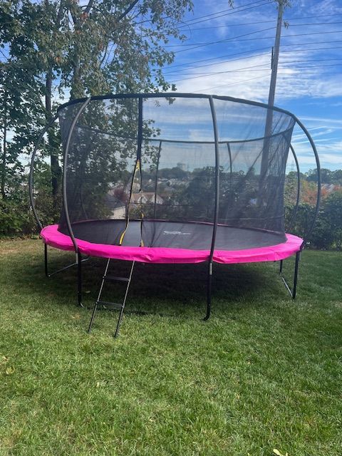 A pink and black trampoline with safety net sits on green grass under a blue sky.
