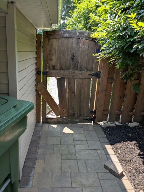 A weathered wooden gate with broken plank, set in a narrow paved walkway next to a building and greenery.