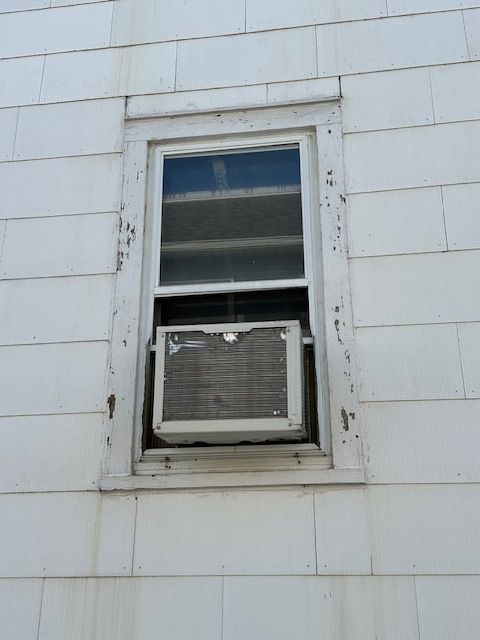 White house exterior with window-mounted air conditioner unit. White siding and window frame.