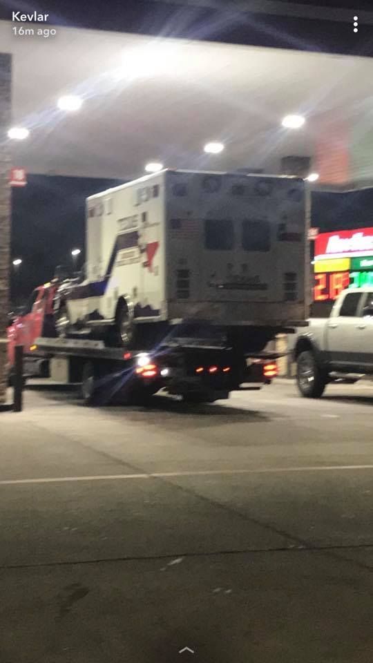An ambulance is being towed by a tow truck at a gas station.
