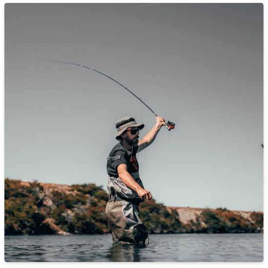 Person fly fishing in a river, holding up a fishing rod. He wears waders and a hat.