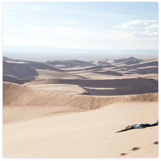 Person lying on sand dune; desert landscape, tan sand, blue sky.