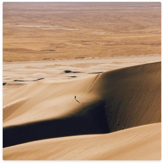 Person walking on a large sand dune, a vast desert landscape in the background, lit by sunlight.