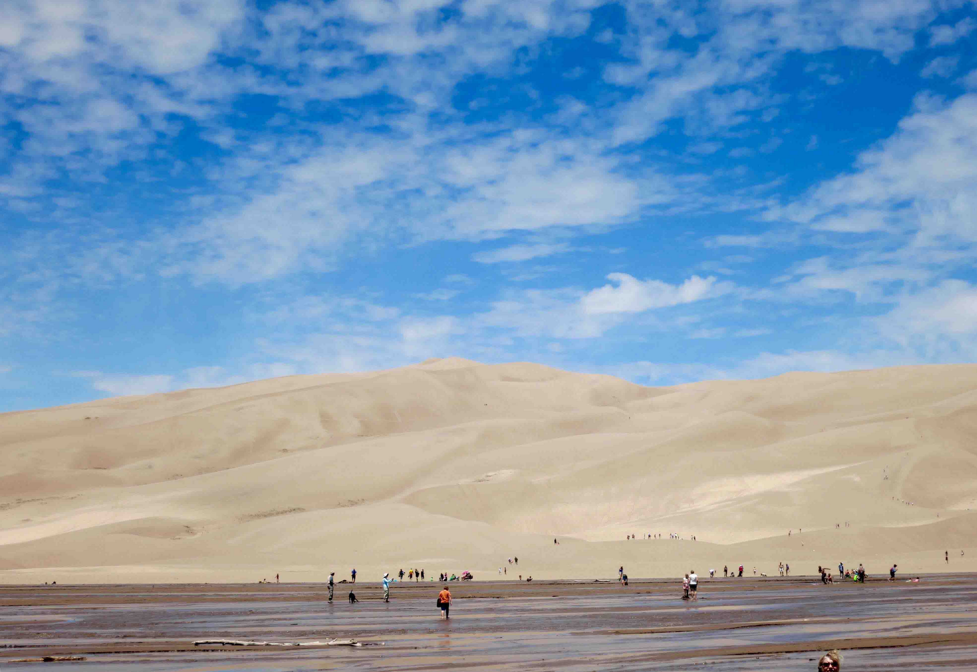 Sandy dunes under a bright blue sky with scattered clouds, people walking in the foreground.
