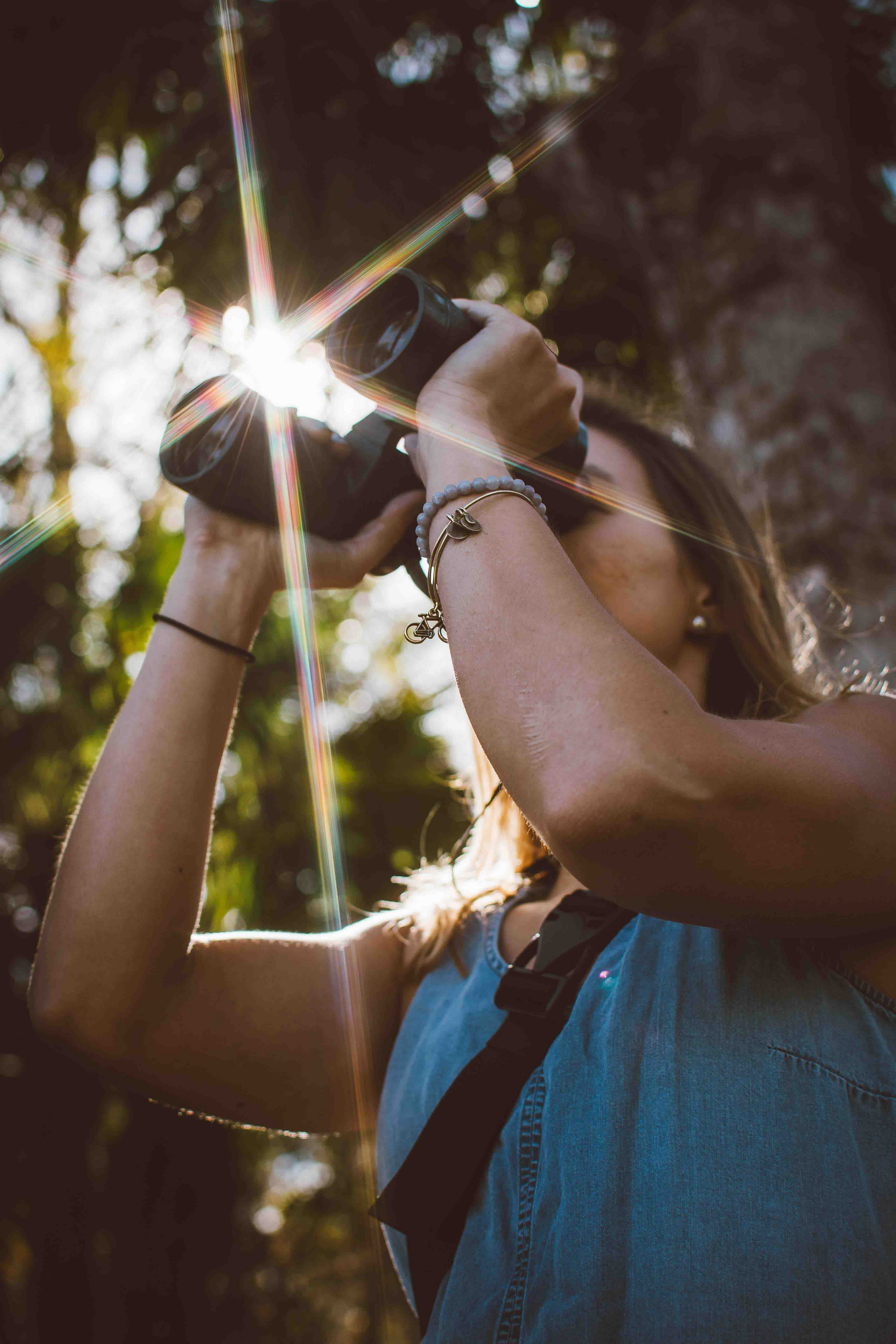 Woman taking a photo with a camera; sunlight creates a starburst effect. She's wearing a denim top and bracelets.