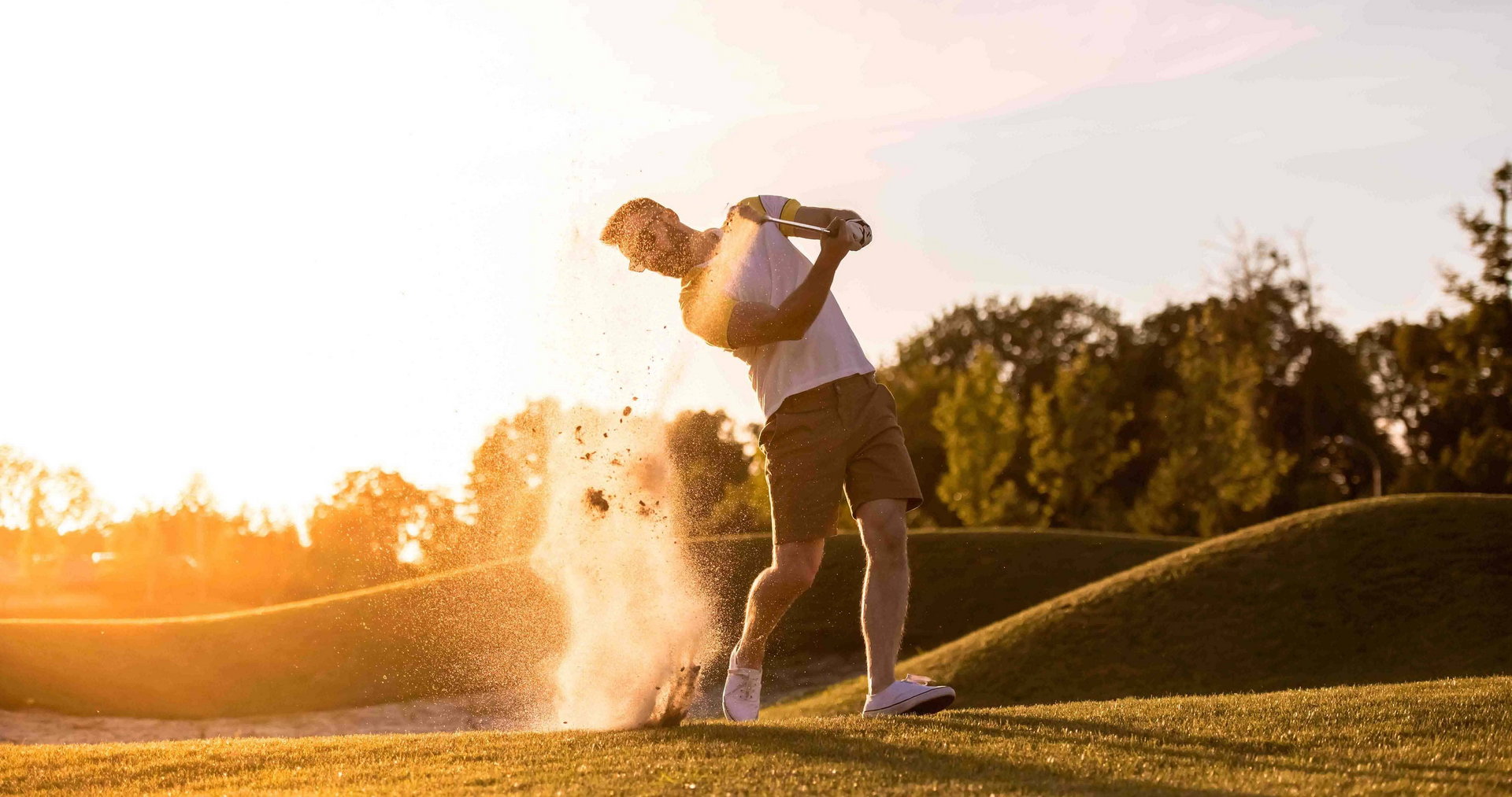 Golfer swinging in a sand trap, kicking up sand, sunny golf course.