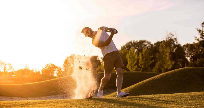 Golfer swinging in a sand trap, kicking up sand, sunny golf course.