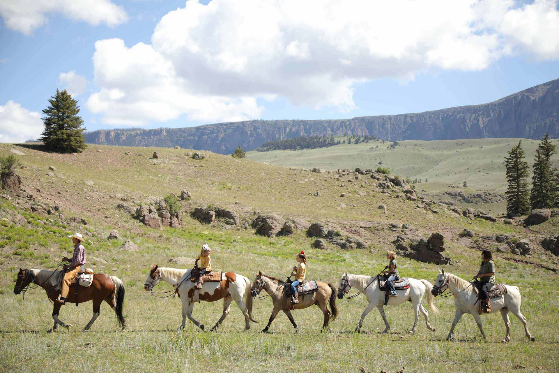 People on horseback ride through a grassy field with mountains in the background.