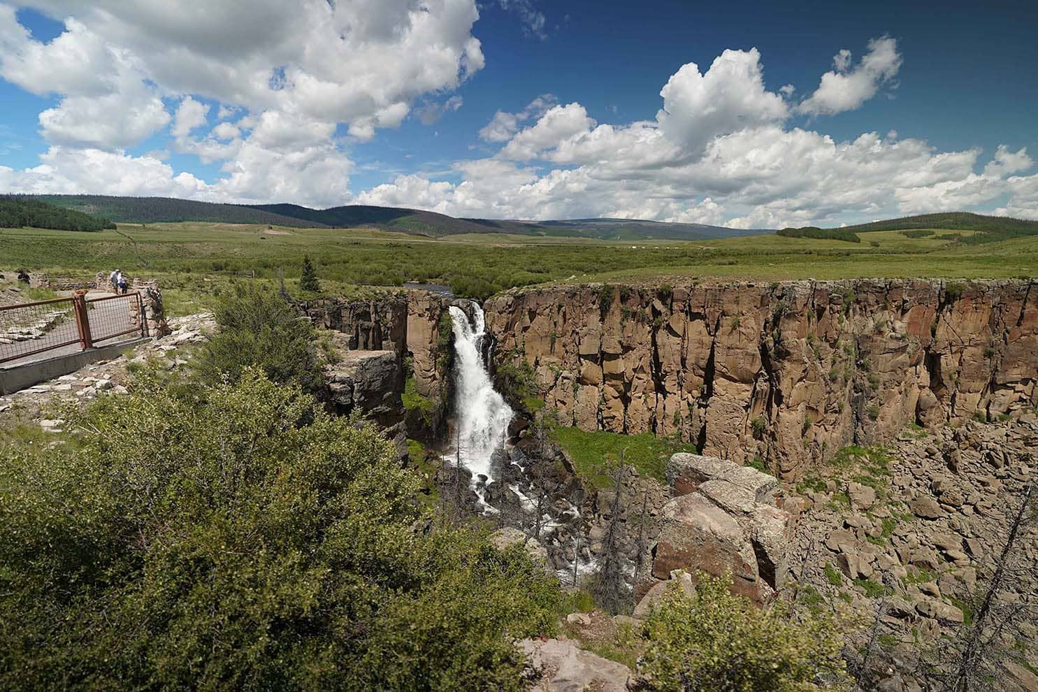 Waterfall cascading into a deep canyon with green foliage, under a blue sky with fluffy clouds.