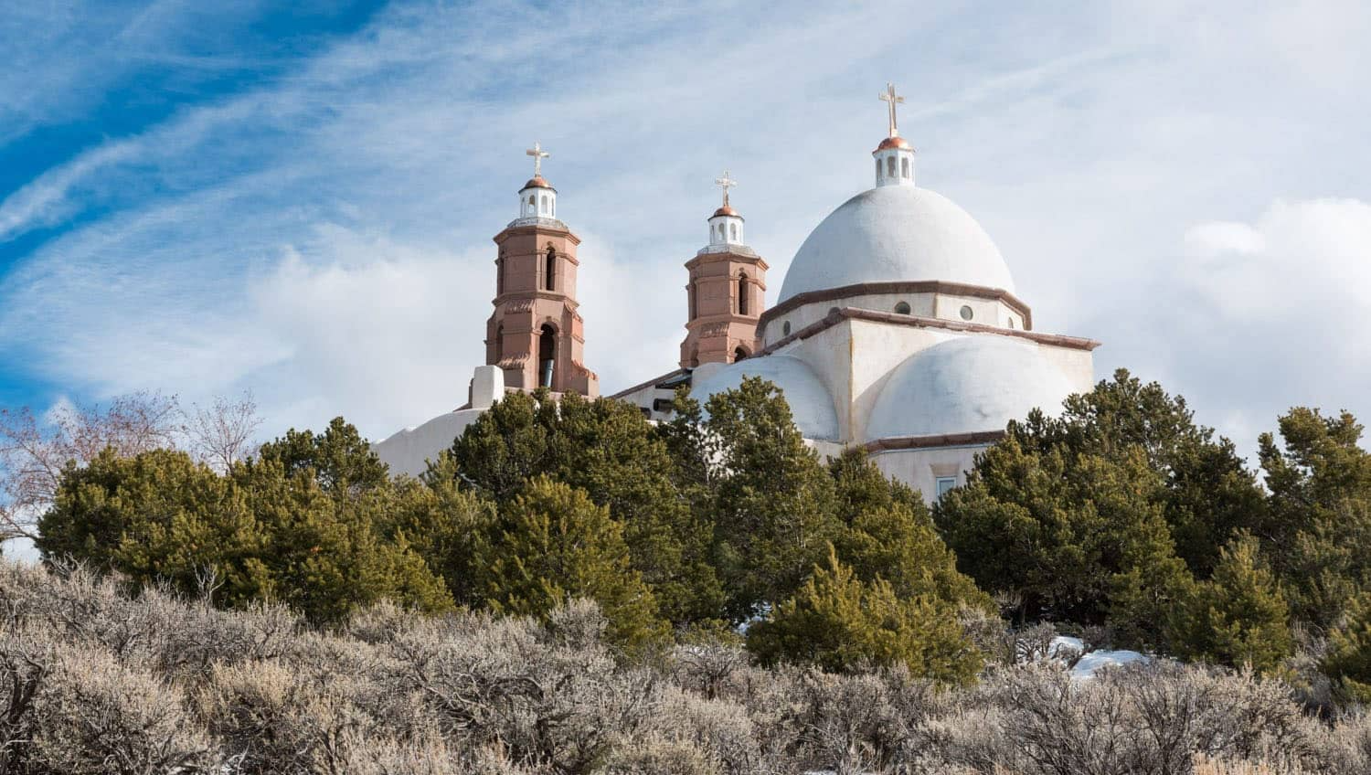 White church with dome and twin bell towers on a hillside, against a cloudy blue sky.