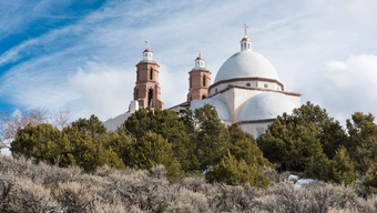 White church with dome and twin bell towers on a hillside, against a cloudy blue sky.