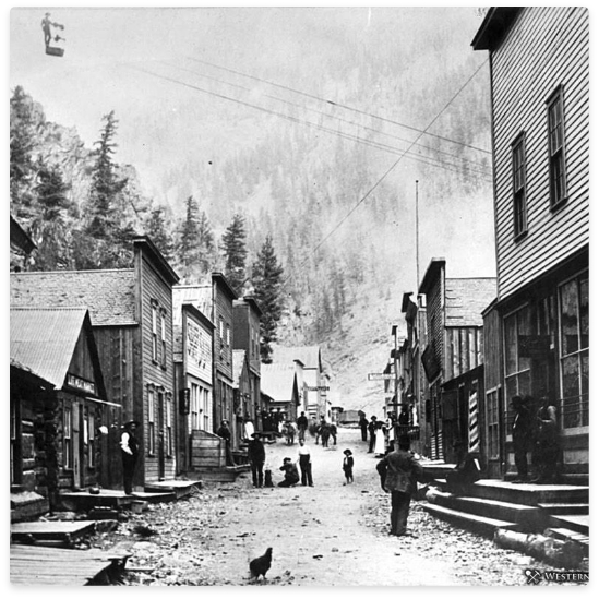 Street in a historic mining town with wooden buildings, people, and a cable car line. Mountains in background.