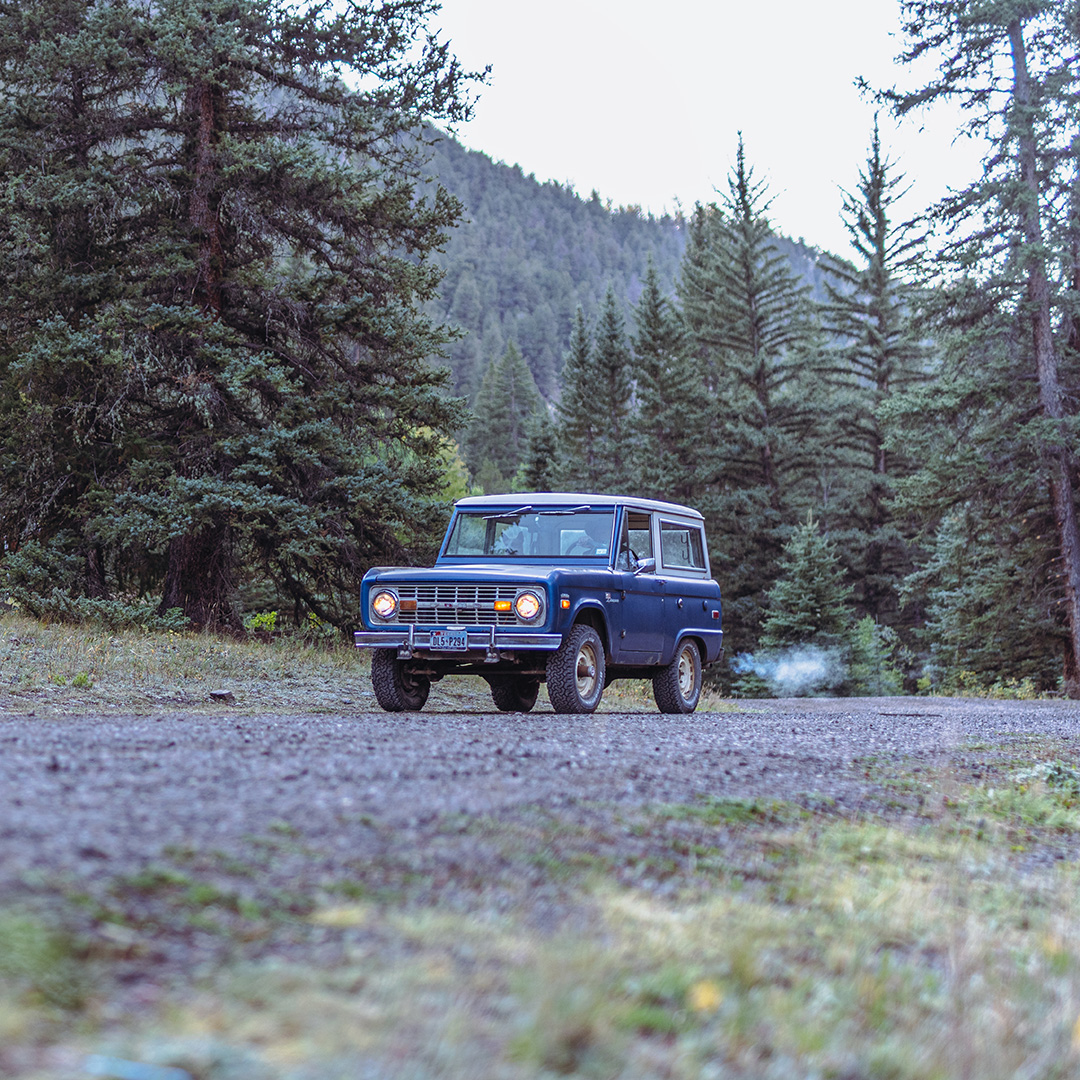 Blue vintage SUV on a dirt road in a forest with mountains in the background.