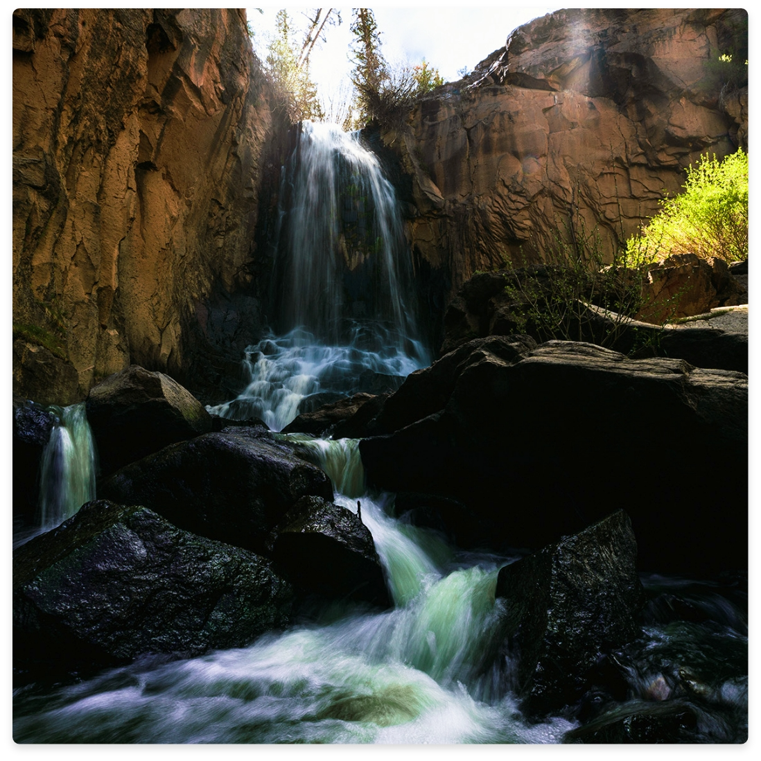 Waterfall cascading over dark rocks, sunlight beams through the top of the cliffs.