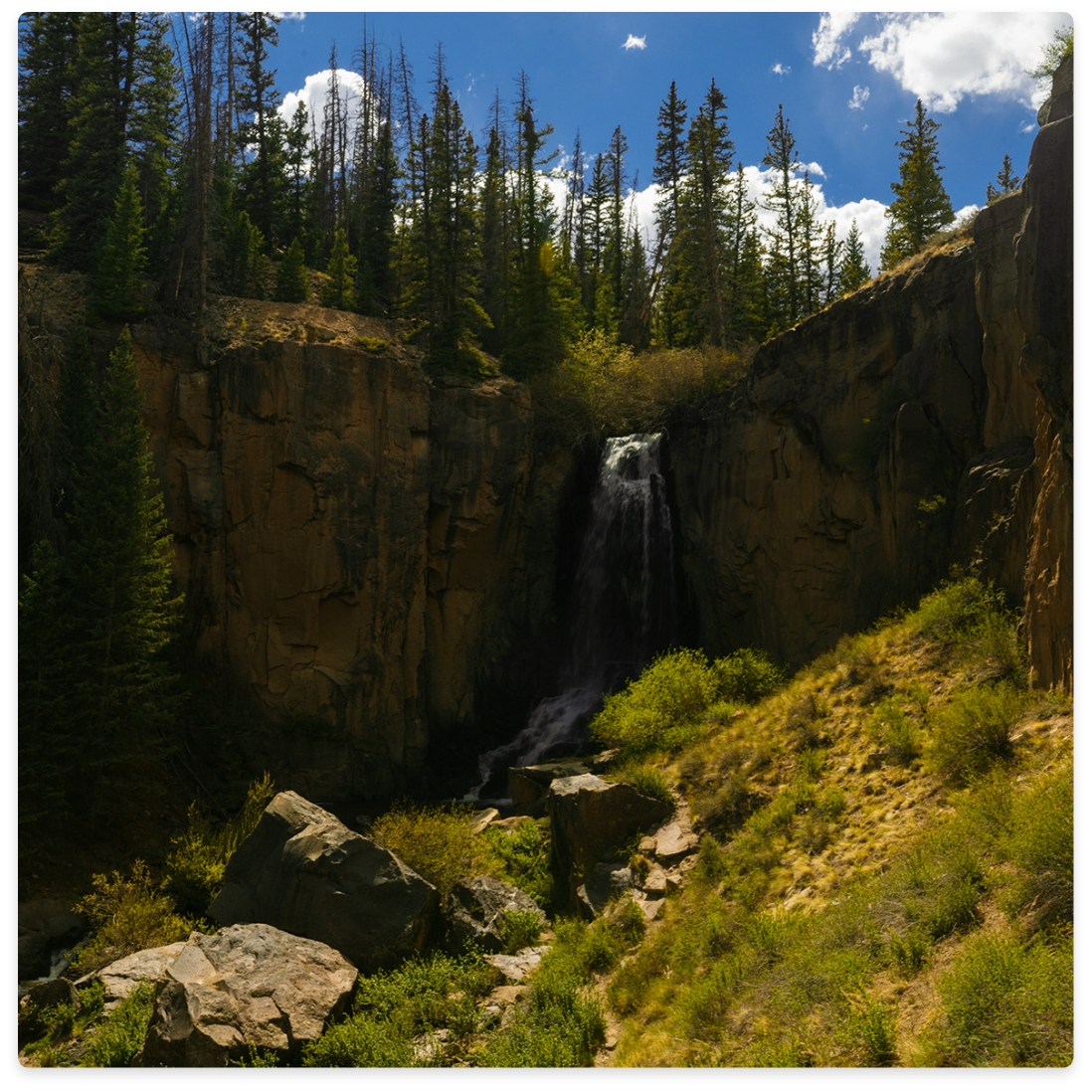 Waterfall cascading down a cliff, surrounded by green grass, rocks, and pine trees under a blue sky.