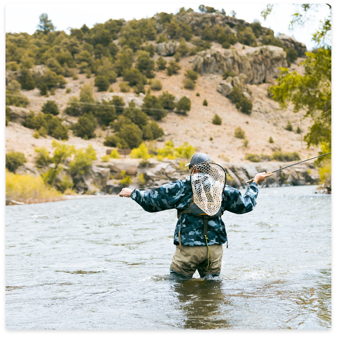 Person fly-fishing in a river, wearing camouflage jacket and waders, with a net on their back. Mountains in the background.