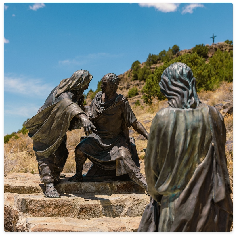 Bronze statues of three figures outdoors, one seated, two standing, hillside background.