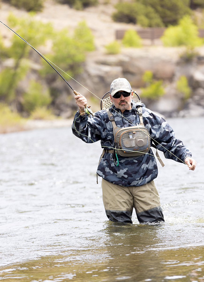 Person fly fishing in a river, wearing waders and camouflage jacket, holding fishing rod, outdoors.