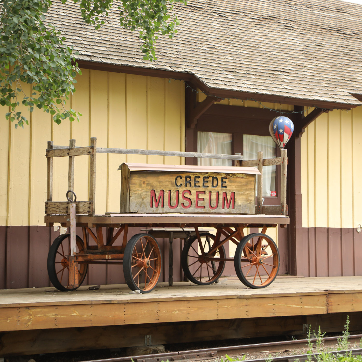Antique wagon with "Creede Museum" sign on a wooden platform in front of a yellow and brown building.
