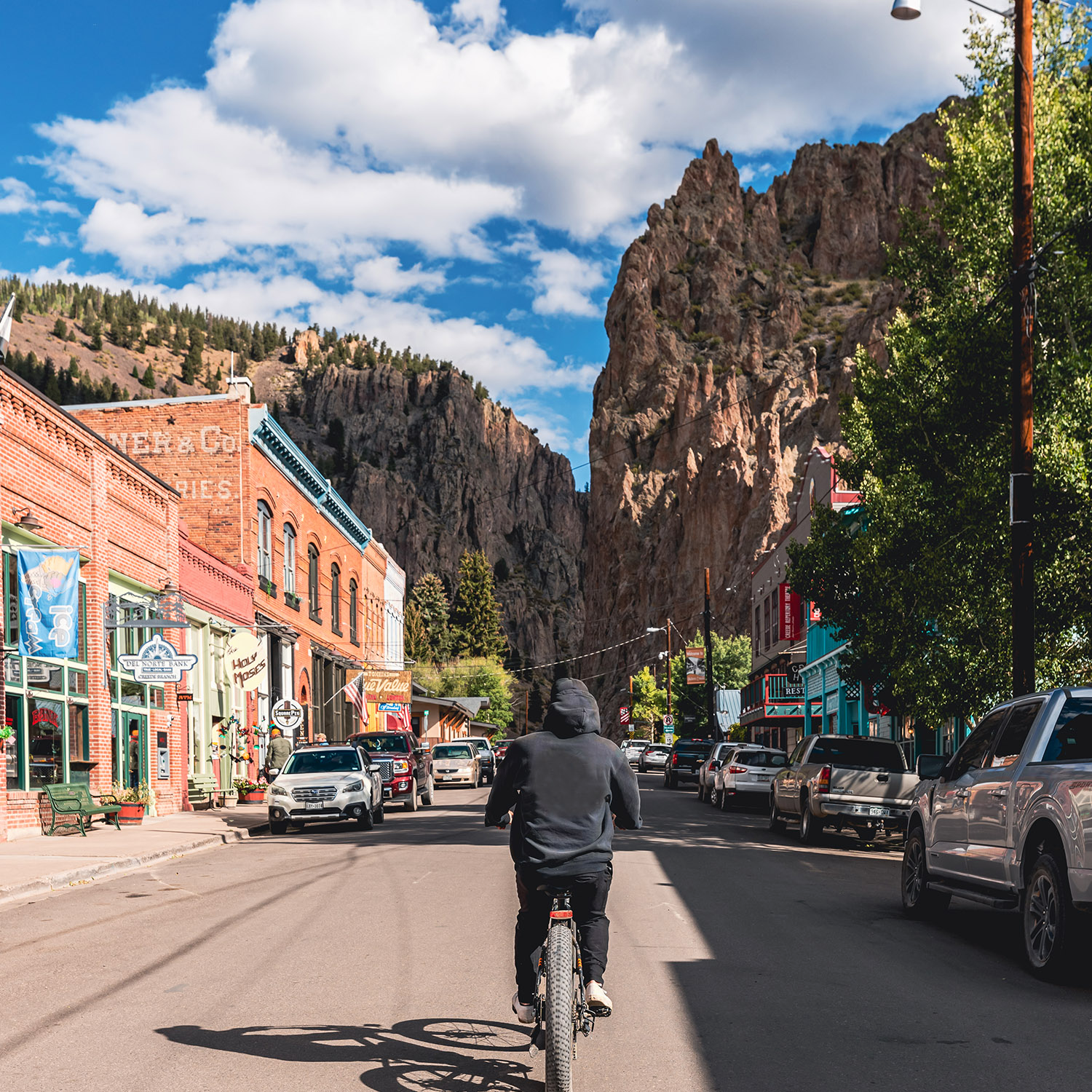 Person biking down a street lined with colorful buildings and mountains in the background. Bright blue sky.