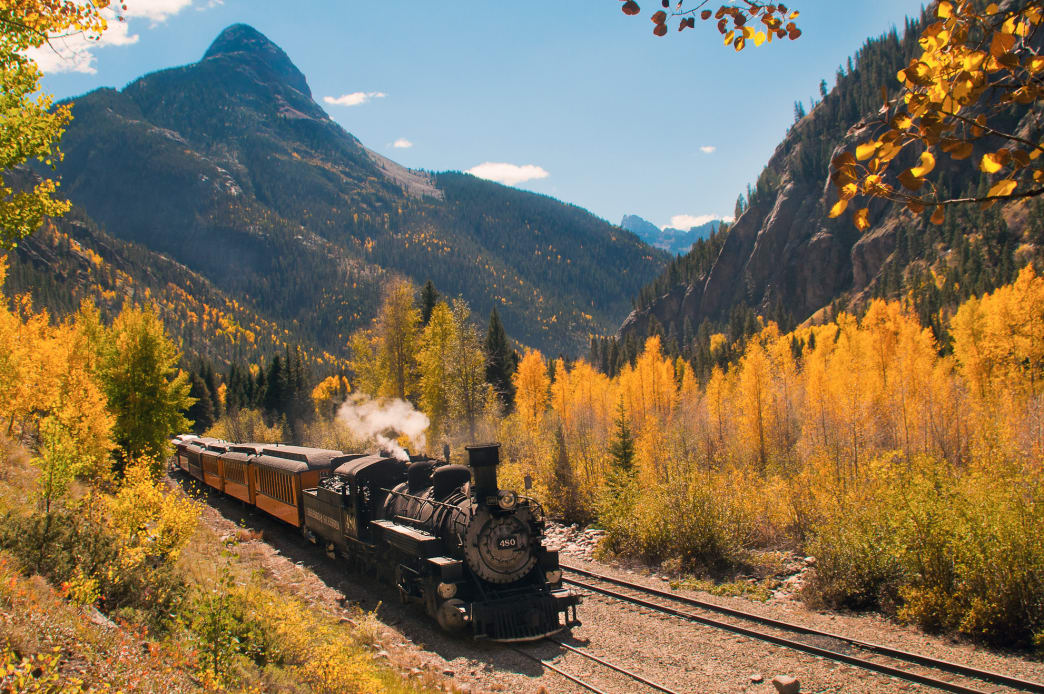 Steam train travels through a mountain pass with fall foliage.