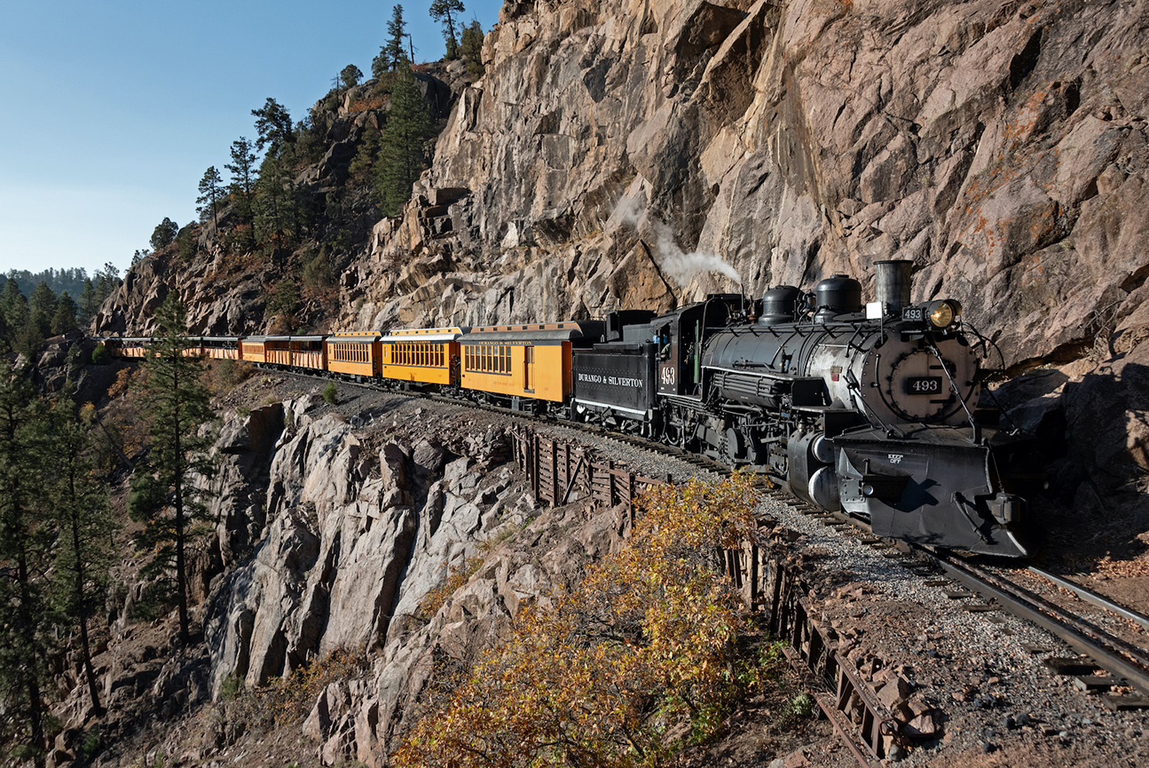 Steam train traveling along a cliffside track, passing orange passenger cars, with rocky mountains visible.