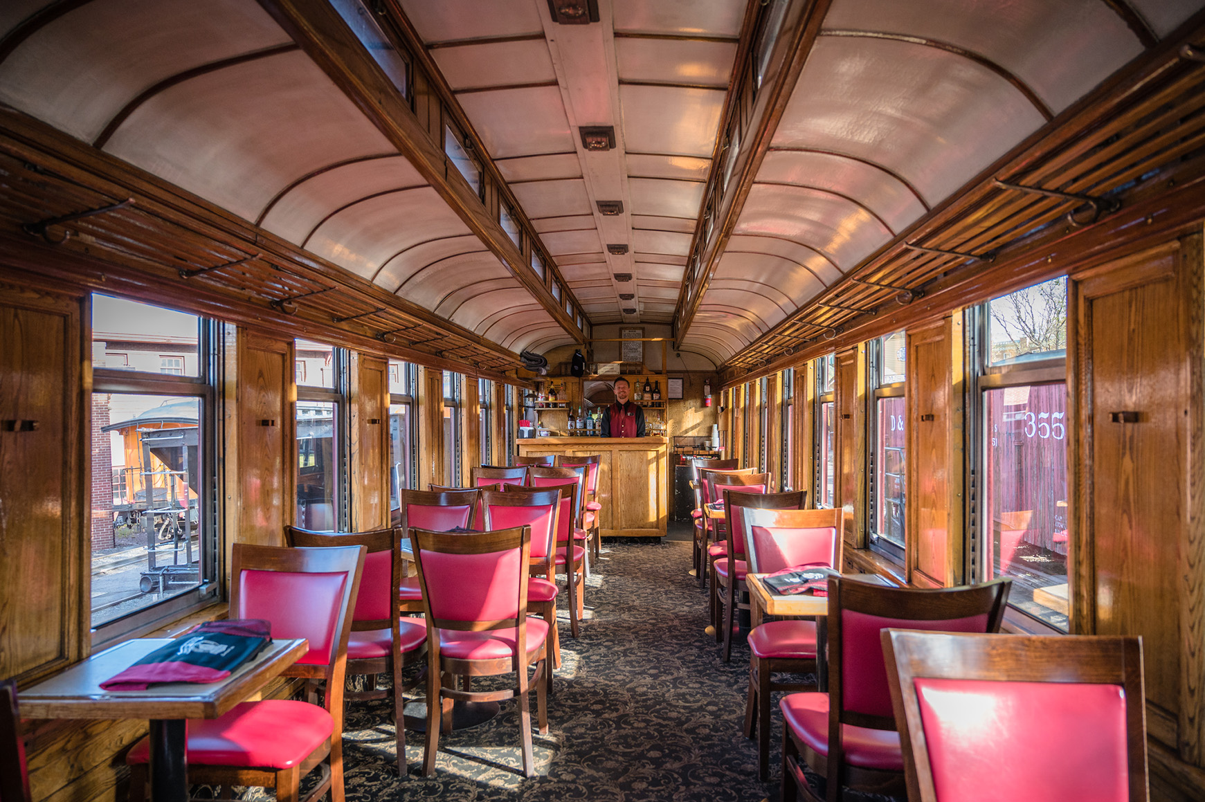 Interior of a vintage train car restaurant with red chairs, tables, and a wooden bar in the distance.