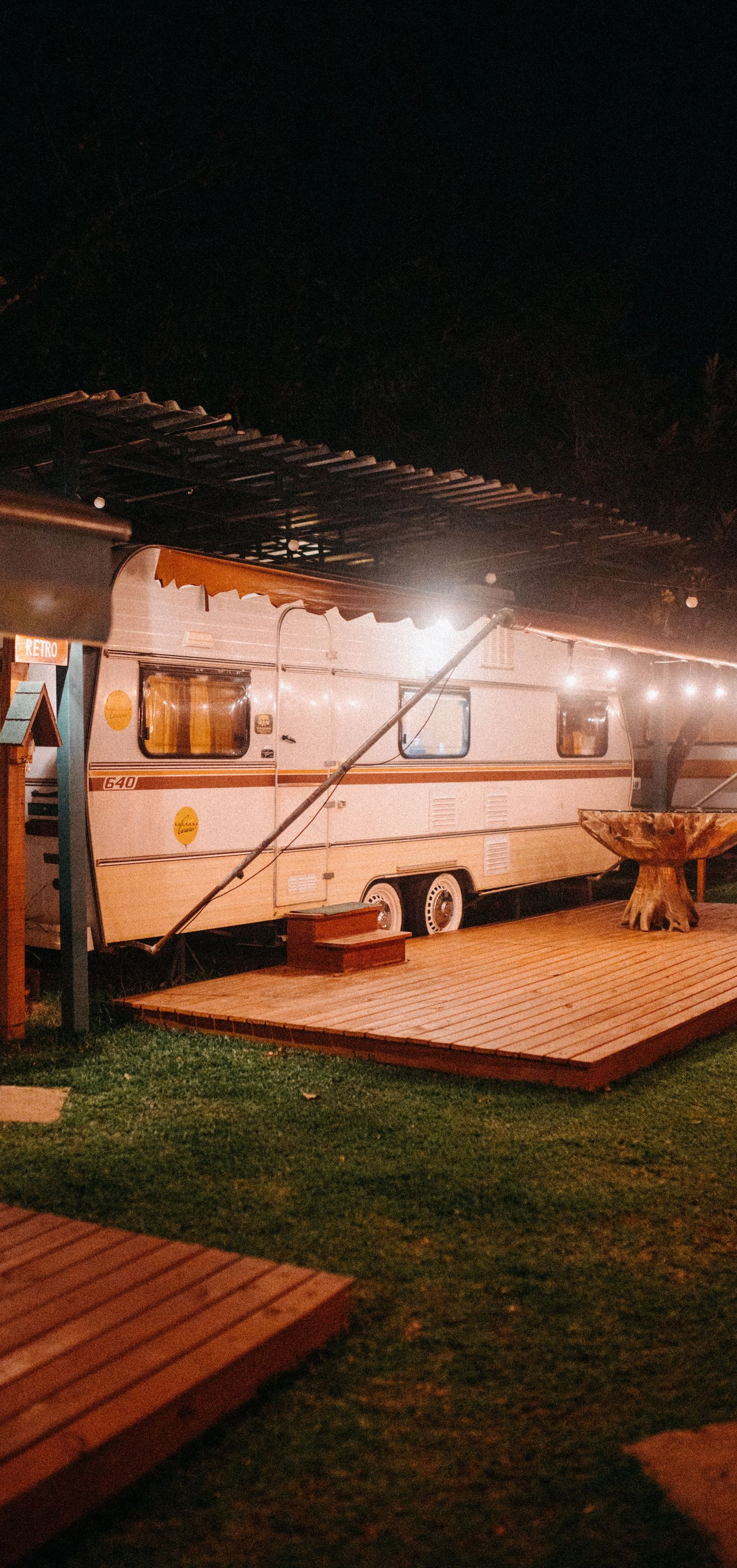 Lit-up white camper trailer at night on a grassy site with wooden decks and string lights