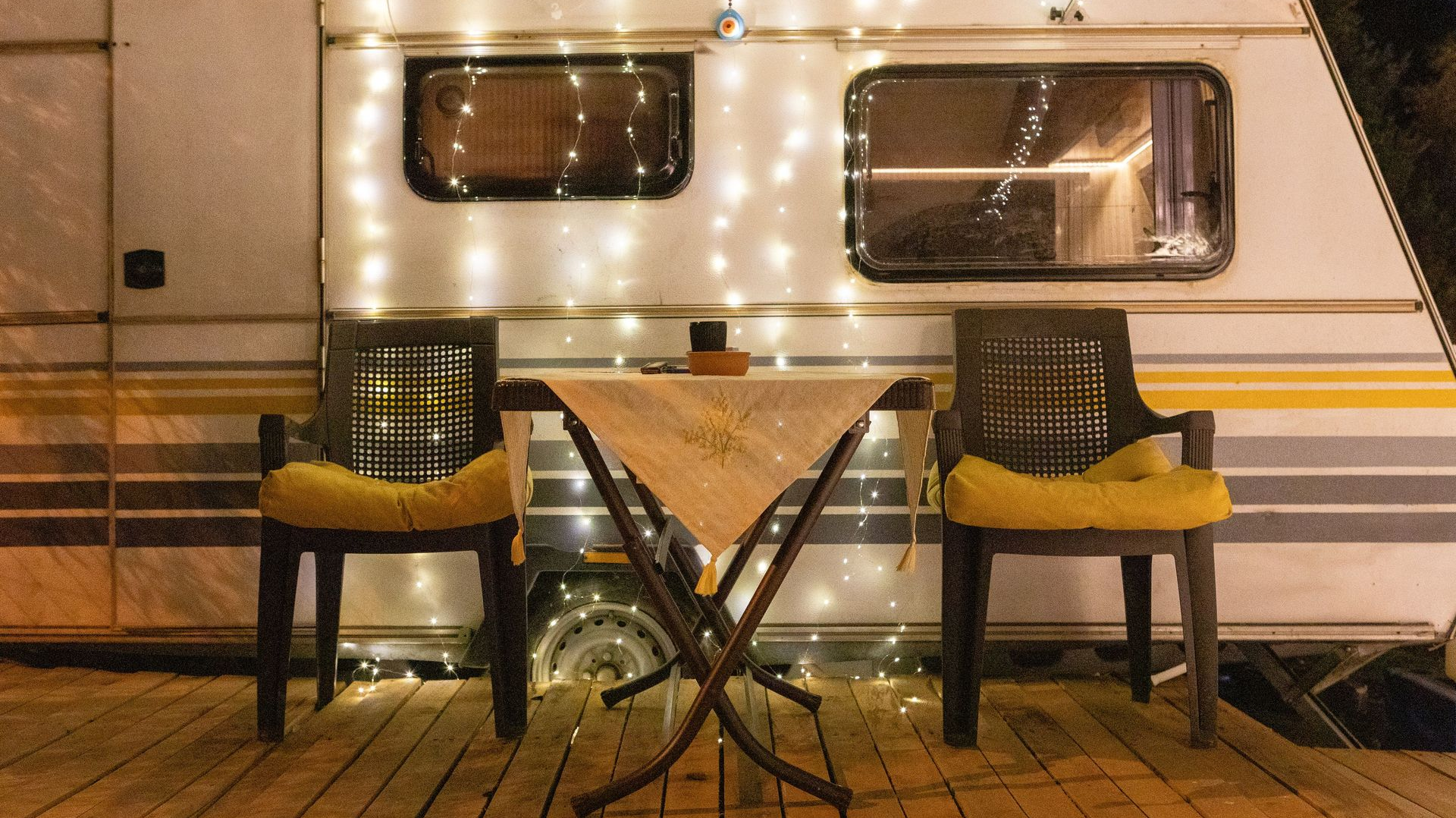 Outdoor café table with two plaid chairs on a wooden deck beside a lit camper at night