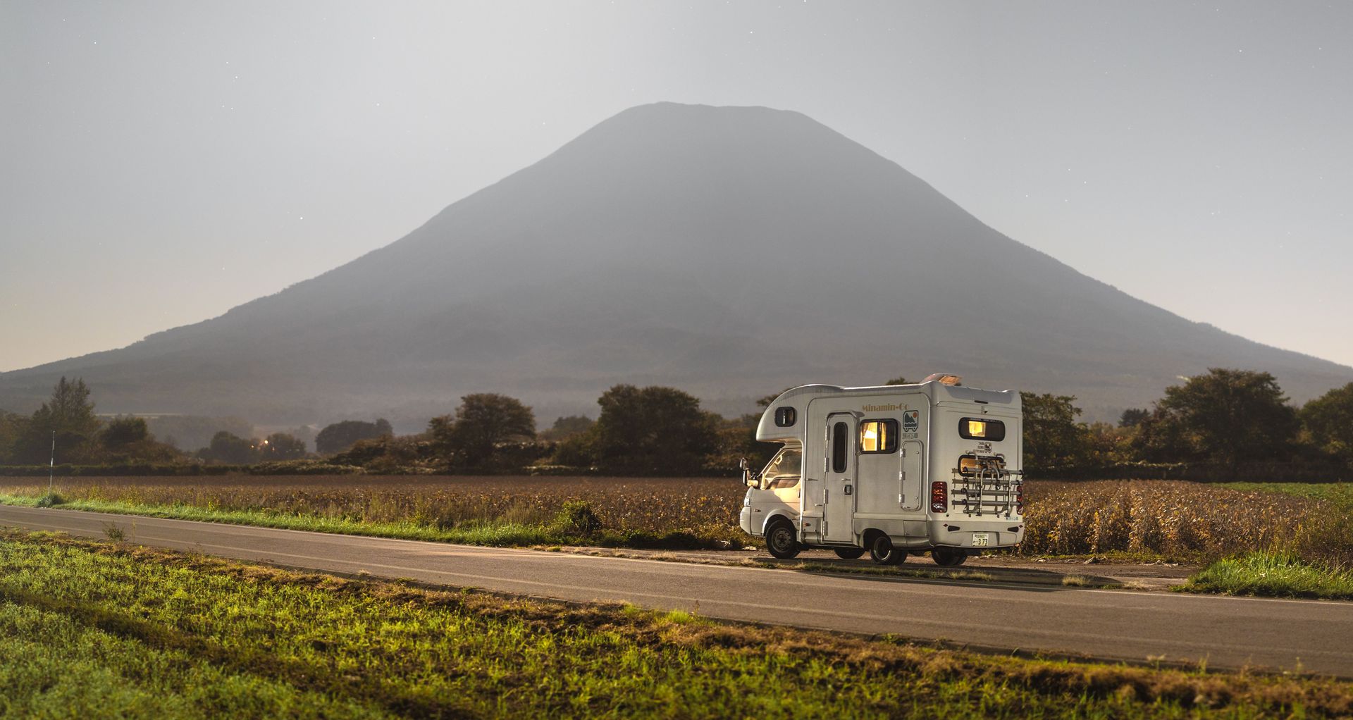 White RV on a rural road with fields, a cloudy sky, and a large mountain in the background