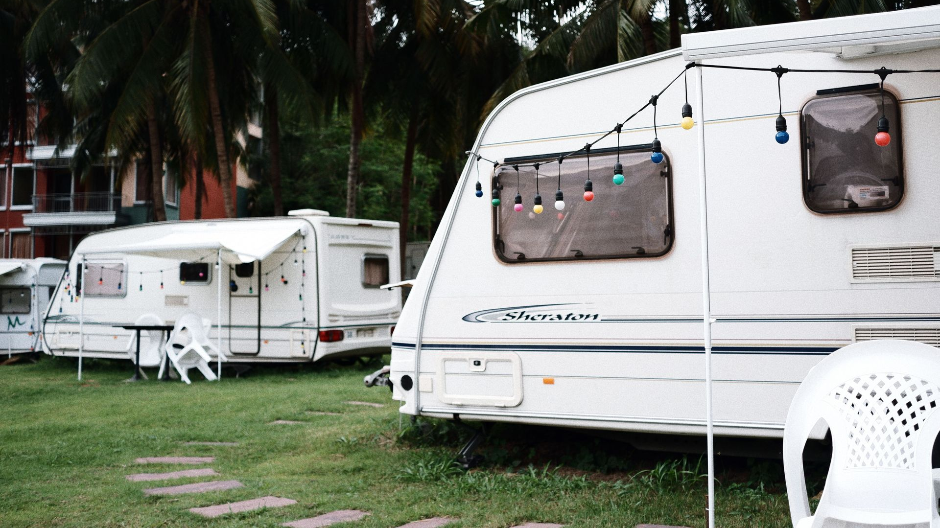 White caravans parked on grass under palm trees, with string lights and outdoor chairs.