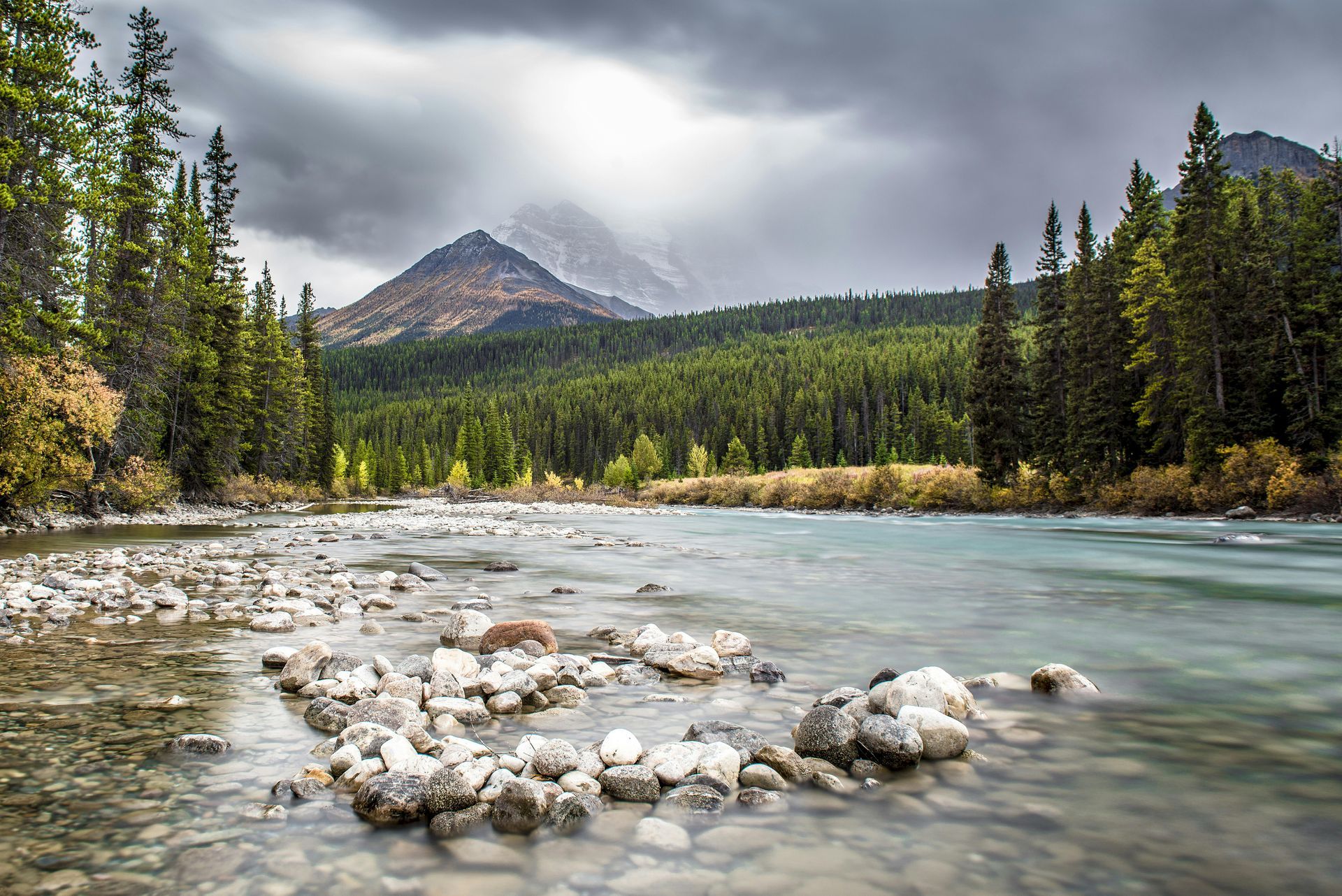 River flows through a rocky foreground, forest, and mountain under cloudy sky.