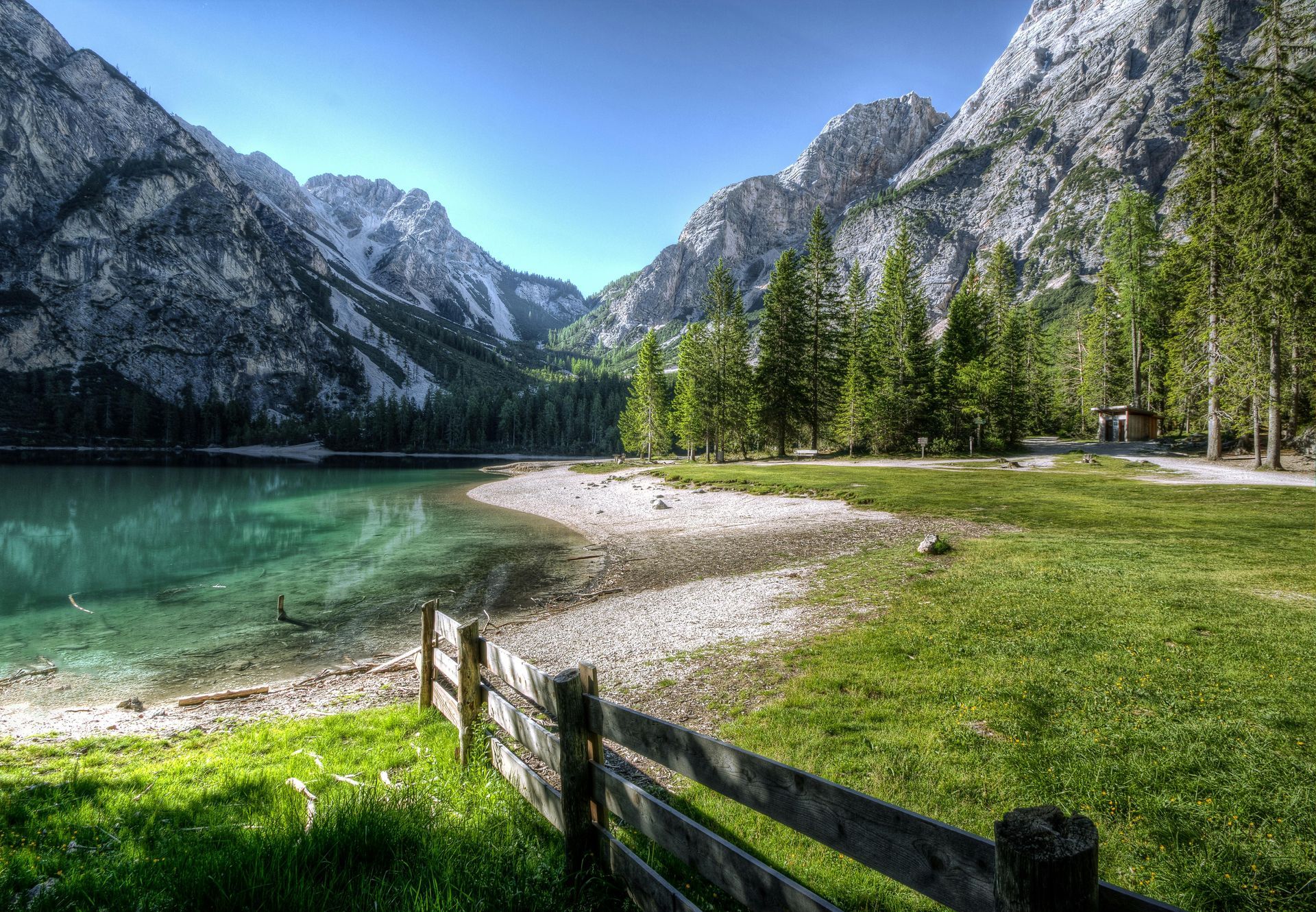 Lush green landscape with lake, mountains, and trees under a clear blue sky. A wooden fence is in the foreground.
