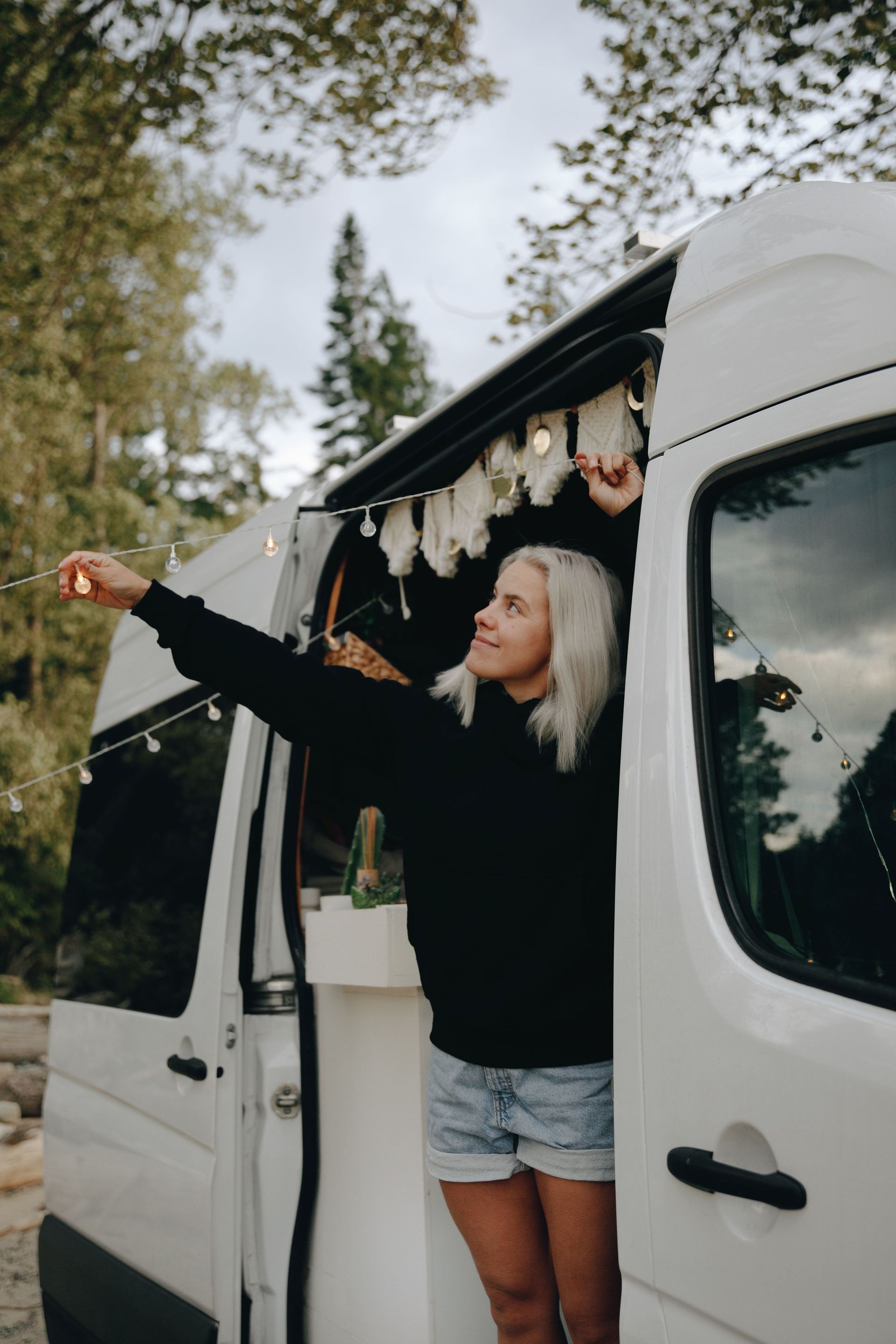 Woman leaning from white van doorway, pointing outside in a wooded setting with hanging decorations.