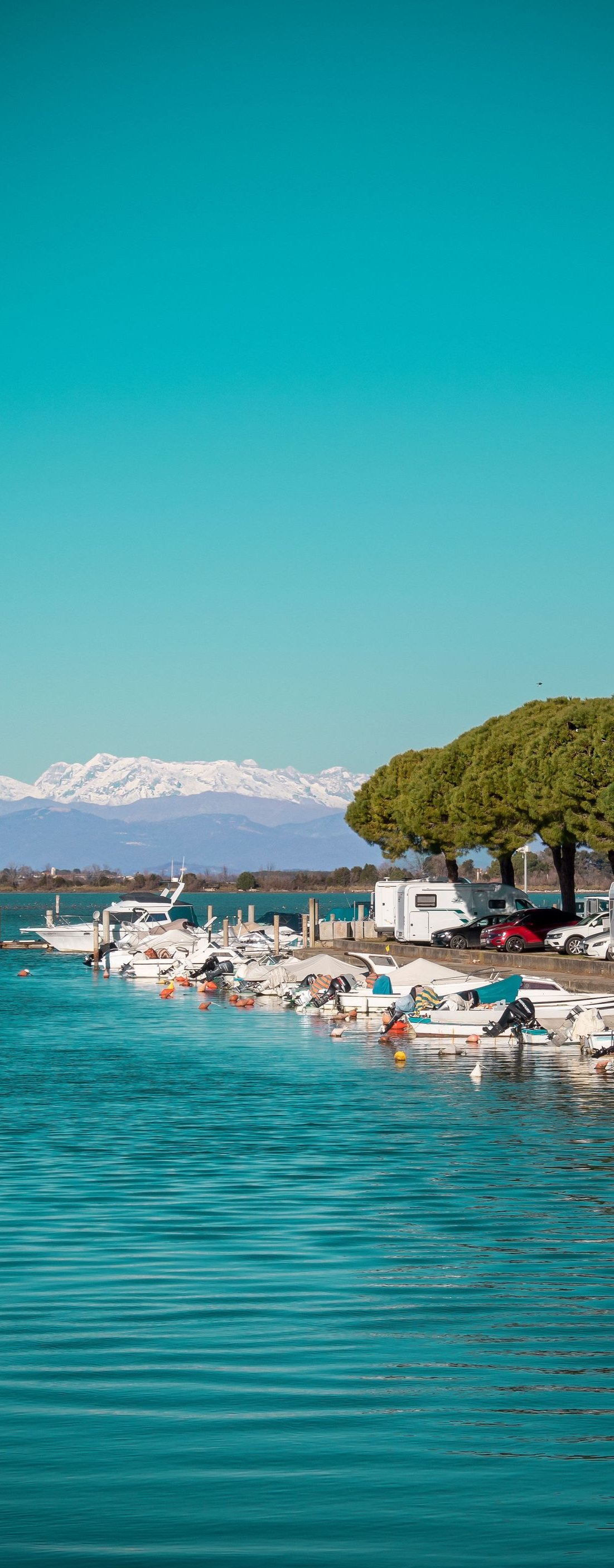 Turquoise sea beside a crowded beach with umbrellas, boats, and snow-capped mountains under a clear blue sky