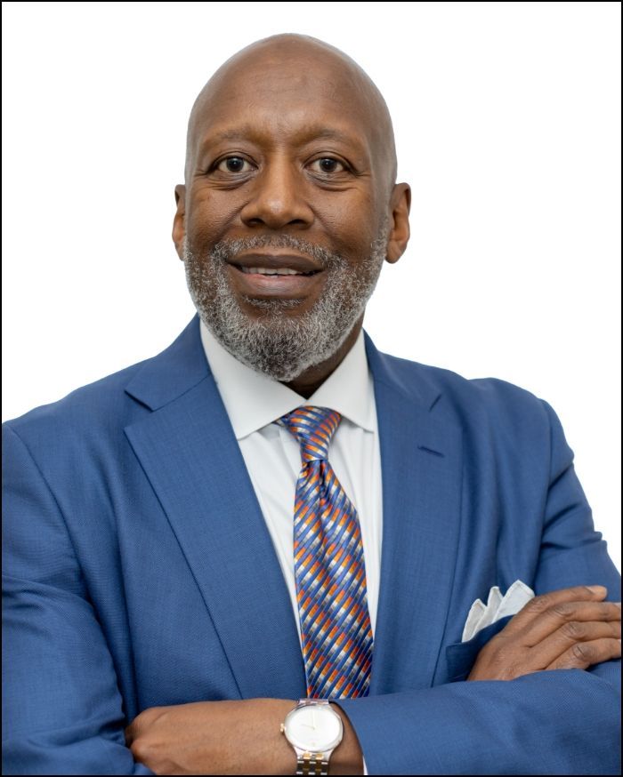 Man in a blue suit with crossed arms, smiling, wearing a patterned tie and watch.