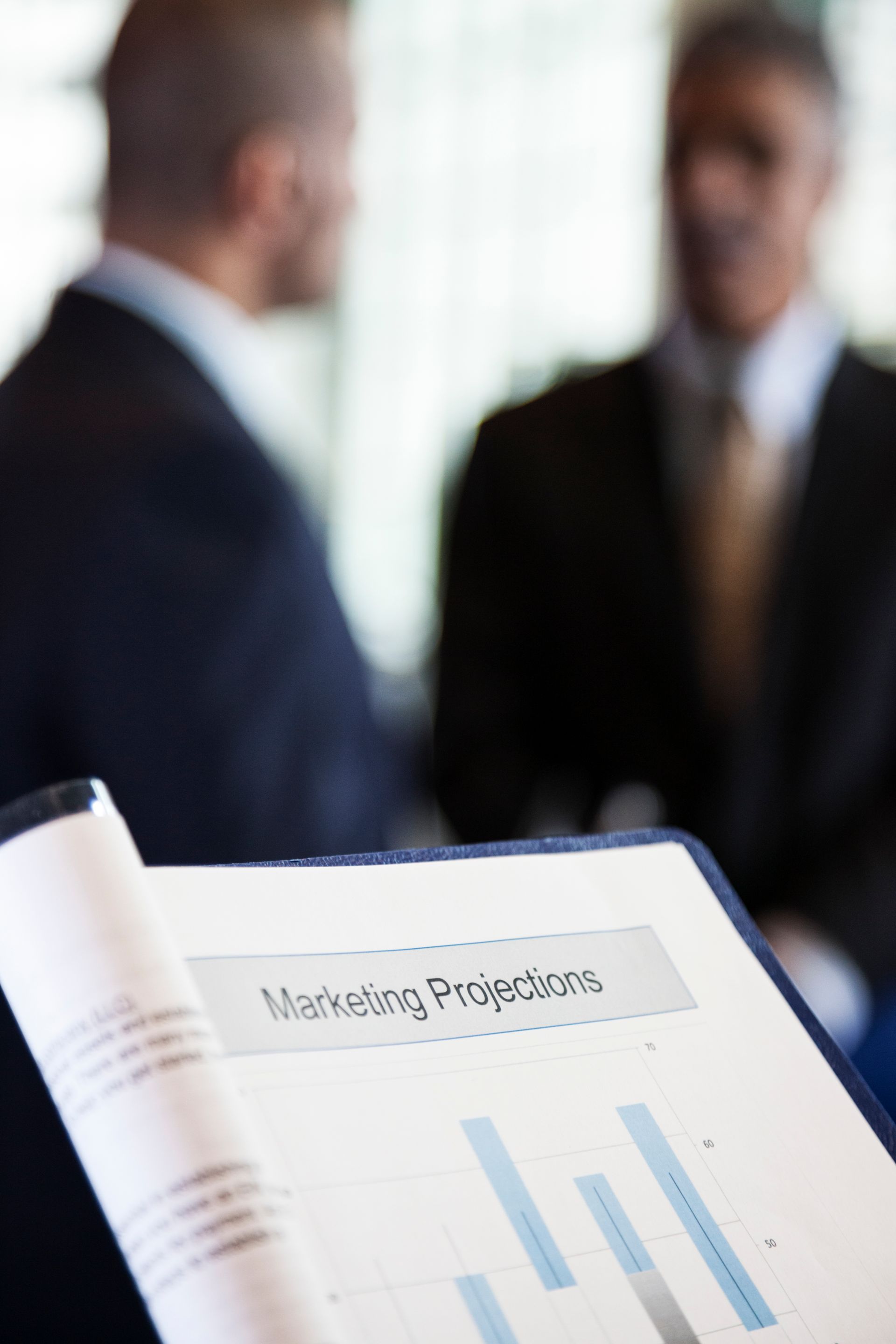 Two businessmen discuss marketing projections, with a document in the foreground.