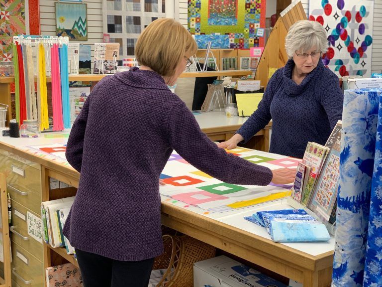 Two women in a quilt shop examining a quilt on a counter. Fabric rolls, books, and quilting supplies are visible.