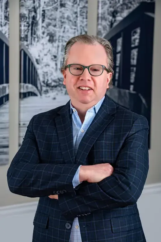 A man in a suit and glasses stands in front of a bookshelf full of books
