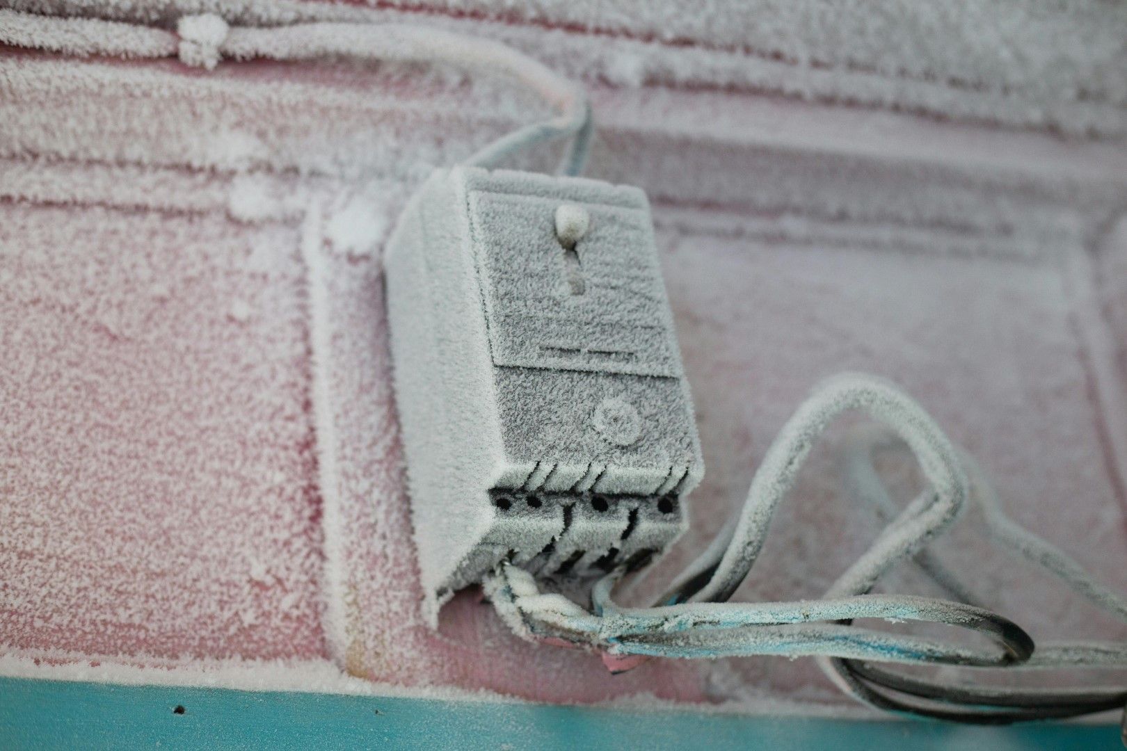 Frost-covered electrical outlet and cord on a pink wall, with snow buildup around the socket.