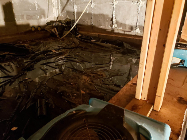 Basement floor covered with black plastic sheeting and debris near exposed wooden framing.