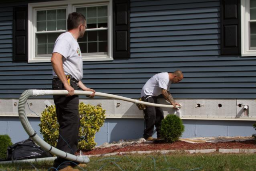 Two workers operating hoses near a blue house exterior, one standing and one crouched by the foundation.
